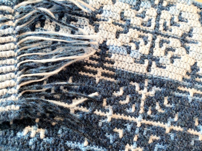 May include: Close-up of a handmade crochet blanket with a geometric pattern in shades of blue and white. The blanket has a fringe of white yarn along one edge. The design features a repeating square motif.