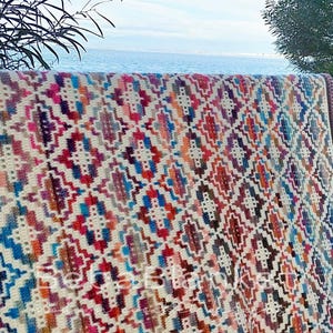 May include: A colorful, geometric patterned blanket with a cream background. The blanket features a repeating design in shades of blue, red, orange, purple, and brown. The blanket is draped over a railing with a view of the ocean in the background.