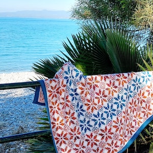 May include: A colorful crocheted blanket with a geometric pattern in shades of orange, blue, and white. The blanket is draped over a railing overlooking a beach.