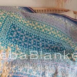 Puede incluir: Una manta multicolor con un patrón geométrico en tonos azules, turquesas, amarillos y lavanda está sobre una silla de madera. La manta presenta un diseño repetitivo de cuadrados y diamantes. El texto "BebaBlanket" es visible.