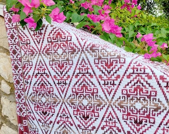 May include: A white and red crocheted throw blanket with a geometric pattern. The blanket is draped over a stone wall with pink flowers in the background.