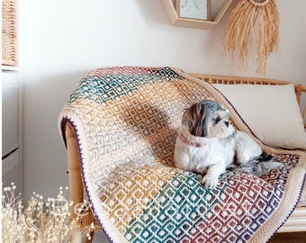 May include: A small dog with brown and white fur is lying on a multi-colored crocheted throw blanket. The blanket has a geometric pattern with shades of green, brown, yellow, and white.