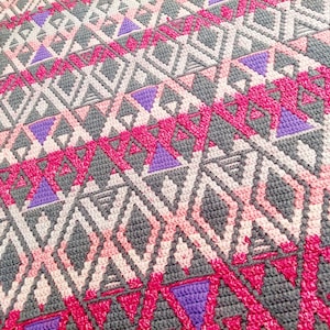 Op de afbeelding: Een gehaakt deken met een geometrisch patroon in tinten roze, paars en grijs.