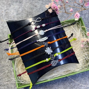 Könnte beinhalten: Ein Set mit 10 bunten Armbändern aus Schnur mit silbernen Anhängern. Die Anhänger umfassen ein Unendlichkeitszeichen, einen Marienkäfer, ein vierblättriges Kleeblatt, einen Schmetterling, eine Feder, eine Blume und eine Perle.