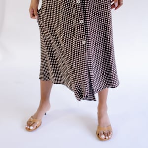 Könnte beinhalten: Eine Frau trägt einen schwarzen Cardigan mit einem braunen und weißen Gingham-Rock. Der Rock hat einen Knopfleisten vorne und eine leicht ausgestellte Silhouette.
