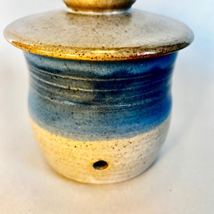 Pode incluir: Um pote de cerâmica com tampa, com um esmalte azul e branco com um acabamento salpicado. O pote tem um pequeno furo no fundo.