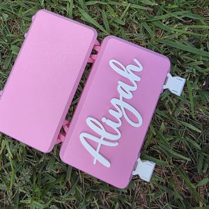 May include: A rectangular pink plastic storage case with a hinged lid and two white side buckles. The top of the case features the name Aliyah in raised white script lettering. The case has a smooth matte finish and a simple functional design.