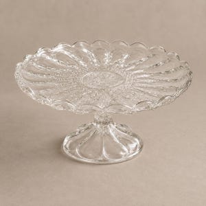 May include: A clear glass cake stand featuring a circular plate with a scalloped edge and a radial pattern of embossed petals. The stand is supported by a short, wide, fluted pedestal base that matches the petal design on the plate surface.