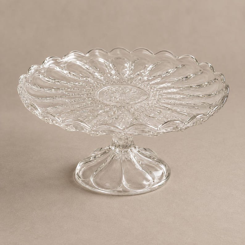 May include: A clear glass cake stand featuring a circular plate with a scalloped edge and a radial pattern of embossed petals. The stand is supported by a short, wide, fluted pedestal base that matches the petal design on the plate surface.