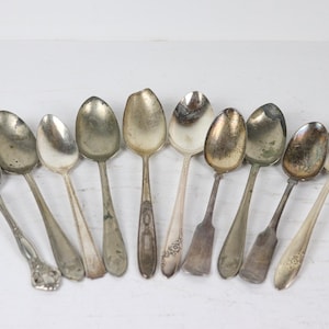 May include: An assortment of antique silver spoons, each with a unique design and patina. The spoons vary in size and shape, with some featuring ornate handles and others with simpler designs. The spoons are arranged on a white surface.