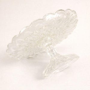 May include: A clear glass cake stand featuring a circular plate with a scalloped edge and a textured, pressed pattern. The stand is supported by a central pedestal base with a matching pressed glass design, providing a stable elevated platform.