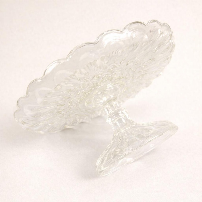 May include: A clear glass cake stand featuring a circular plate with a scalloped edge and a textured, pressed pattern. The stand is supported by a central pedestal base with a matching pressed glass design, providing a stable elevated platform.