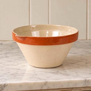 May include: A cream-colored ceramic mixing bowl with a wide, smooth, burnt-orange band around the upper exterior rim. The bowl features a deep, conical shape with a small, integrated pouring spout on one side of the rim and a glossy finish.