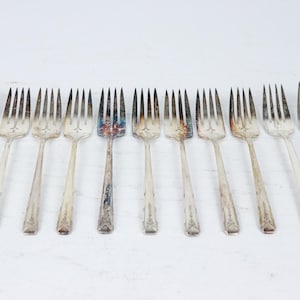May include: A set of antique silver forks displayed in a line. The forks feature tines and long handles with decorative designs. Some forks show signs of age and wear, with tarnishing and discolouration. The forks are set against a white background.