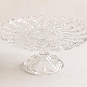 May include: A clear glass cake stand featuring a circular plate with a scalloped edge and a radial pattern of embossed lines extending from the centre. The stand is supported by a short, wide, fluted pedestal base with a rounded, scalloped foot.