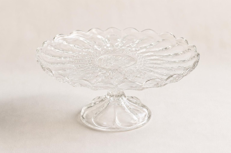 May include: A clear glass cake stand featuring a circular plate with a scalloped edge and a radial pattern of embossed lines extending from the centre. The stand is supported by a short, wide, fluted pedestal base with a rounded, scalloped foot.
