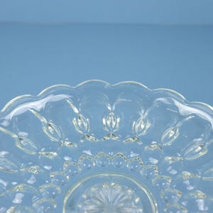 May include: A clear glass plate featuring a scalloped edge and a pressed pattern of raised oval shapes radiating from a central starburst design. The transparent glass has a smooth, reflective surface with a distinct textured geometric motif.