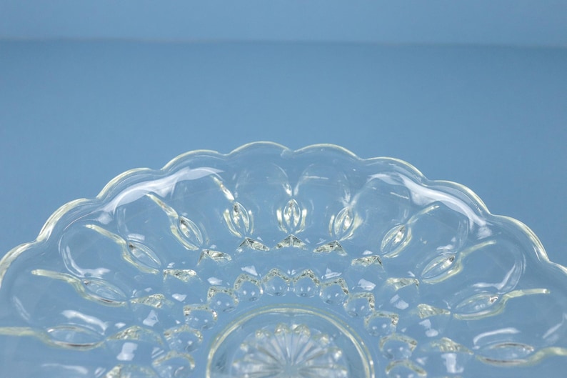 May include: A clear glass plate featuring a scalloped edge and a pressed pattern of raised oval shapes radiating from a central starburst design. The transparent glass has a smooth, reflective surface with a distinct textured geometric motif.