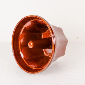 May include: A glossy, dark brown ceramic bundt cake mold featuring a fluted, scalloped interior design with a central tube. The baking pan has a smooth, reflective finish and a wide, circular rim, showcasing a deep, rich color throughout the piece.