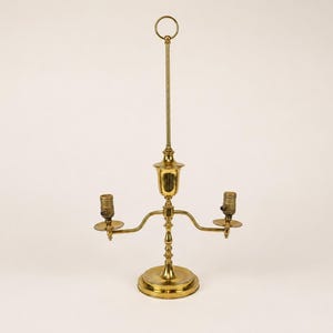 May include: A brass two-light table lamp featuring a central vertical rod topped with a circular ring handle. The lamp has a round base, a central stem with decorative detailing, and two curved arms extending outward, each holding a light socket.