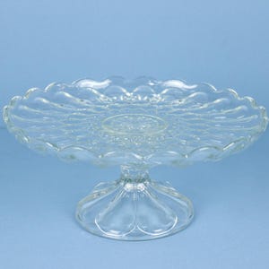 May include: A clear glass cake stand featuring a circular plate with a scalloped edge and a pressed pattern of radiating lines. The stand is supported by a sturdy, wide, circular pedestal base with a smooth, moulded glass construction.