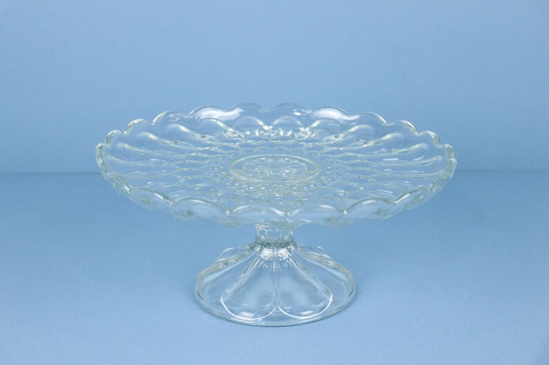 May include: A clear glass cake stand featuring a circular plate with a scalloped edge and a pressed pattern of radiating lines. The stand is supported by a sturdy, wide, circular pedestal base with a smooth, moulded glass construction.