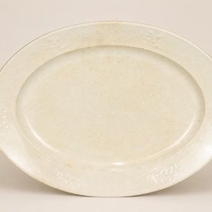 May include: An oval-shaped off-white ceramic platter featuring a smooth, slightly recessed center and a wide, flat rim. The rim is decorated with subtle, embossed floral motifs positioned at intervals around the perimeter of the dish.