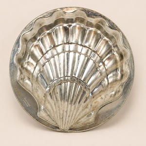 May include: A circular silver-toned metal baking mold featuring a raised, scalloped shell design with radiating ridges. The mold has a smooth, flat outer rim and a central, deeply embossed shell shape. The text "MADE IN FRANCE" is visible.