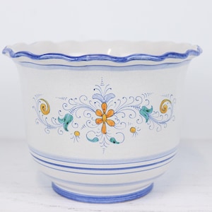 May include: White ceramic planter with a scalloped rim and blue accents. The planter features a central floral design in orange, blue, and yellow. Blue stripes encircle the base. A decorative piece for indoor or outdoor use.