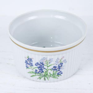 May include: White ceramic ramekin with a gold rim and vertical ribbed design. The ramekin features a floral design of blue and green flowers. Ideal for baking individual portions of desserts or savory dishes.