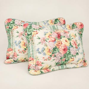 May include: Two rectangular quilted pillow shams featuring a cream-colored background with a vibrant floral pattern of pink, yellow, and red roses, tulips, and green foliage. The edges are finished with a green and white striped border trim.
