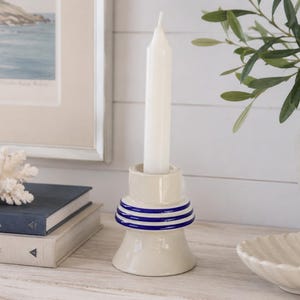 May include: A cream-colored ceramic candle holder with three horizontal blue rings around the center. The holder is cylindrical with a flared base and holds a single white taper candle. The design is smooth and glossy, featuring a simple structure.
