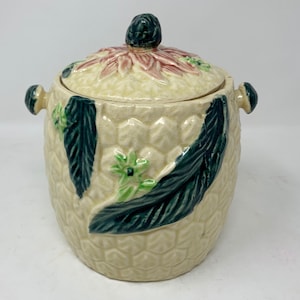 May include: A vintage ceramic cookie jar with a floral design. The jar is white with green leaves and pink flowers. The lid has a raised floral design and a knob in the shape of a flower bud.