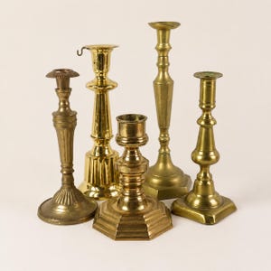 May include: A collection of five brass candlesticks of varying heights and designs. Each candlestick features a metallic gold finish, a central stem, and a base. The shapes include round, octagonal, and square bases with smooth or fluted stems.