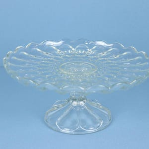 May include: A clear glass cake stand featuring a circular plate with a scalloped edge and a textured, pressed pattern. The stand is supported by a pedestal base with a wide, rounded foot, showcasing a transparent design with reflective surfaces.