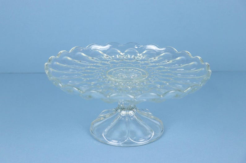May include: A clear glass cake stand featuring a circular plate with a scalloped edge and a textured, pressed pattern. The stand is supported by a pedestal base with a wide, rounded foot, showcasing a transparent design with reflective surfaces.