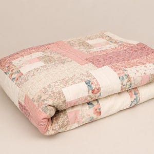 May include: A folded quilt featuring a patchwork design with various floral patterns in shades of pink, cream, and muted blue. The fabric blocks are arranged in a rectangular grid pattern with visible stitching throughout the textile surface.