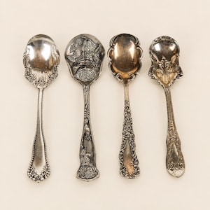May include: A set of four silver-toned metal spoons arranged vertically. Each spoon features a unique handle design with intricate embossed patterns, including floral motifs, scrollwork, and textured edges, with varying bowl shapes and sizes.