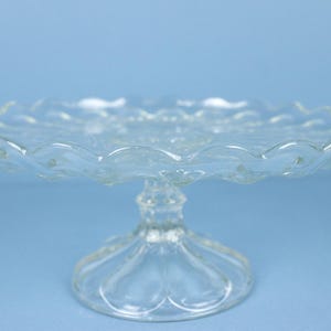 May include: A clear glass cake stand featuring a scalloped edge and a textured pattern on the circular plate. The stand is supported by a central pedestal base with a decorative, faceted design that flares out at the bottom for stability.