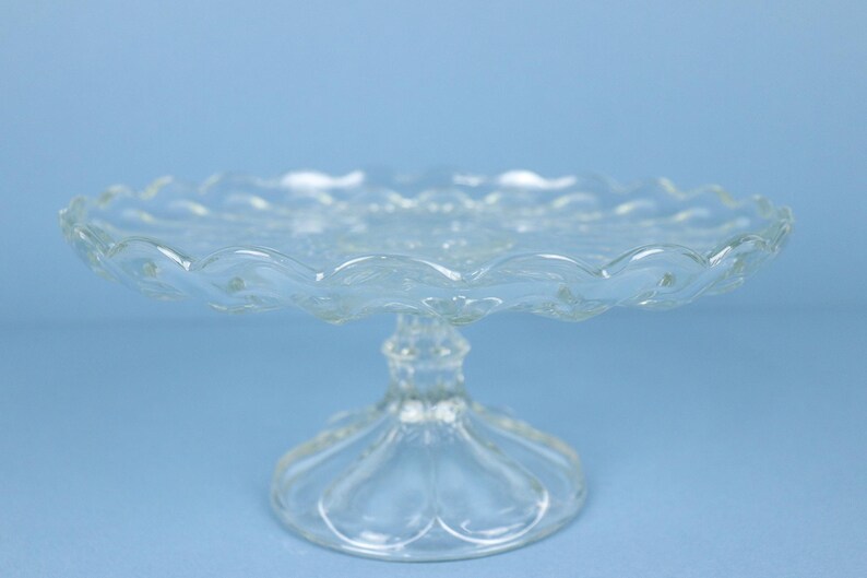 May include: A clear glass cake stand featuring a scalloped edge and a textured pattern on the circular plate. The stand is supported by a central pedestal base with a decorative, faceted design that flares out at the bottom for stability.
