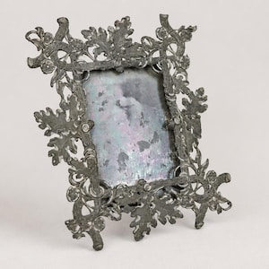 May include: A rectangular picture frame made of dark gray metal with an intricate, raised relief design featuring oak leaves and acorns. The frame surrounds a central, weathered, and mottled glass pane with a faded, indistinct light-colored image.