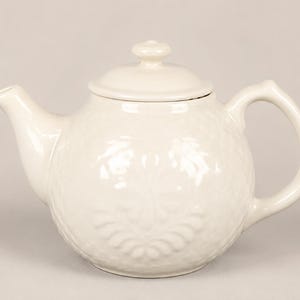 May include: A cream-colored ceramic teapot with a rounded body, a curved spout, and a matching lid with a small knob handle. The exterior surface features a subtle, embossed floral pattern that covers the main body of the teapot.