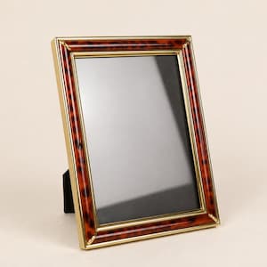 May include: A rectangular picture frame featuring a glossy tortoiseshell pattern border with dark brown and amber spots, accented by thin gold-toned metal trim along the inner and outer edges. The frame includes a black backing stand for display.