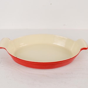 May include: A red and cream-colored oval baking dish with two handles. The dish has a cream-colored interior and a red exterior. The handles are ribbed. This kitchenware is suitable for baking and serving.