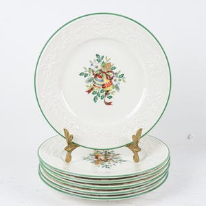 May include: A stack of white ceramic plates with a green border and a floral design. The plates are stacked on a gold-colored plate stand. The top plate is displayed upright, showcasing the full design.