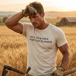 Pode incluir: Uma t-shirt de manga curta cor de creme com o texto preto "OUT OF RANGE FOR THE SUMMER" impresso no peito. A peça é usada por uma pessoa de pele bronzeada, com luvas de trabalho de couro castanho, enquanto segura um cabo de madeira.