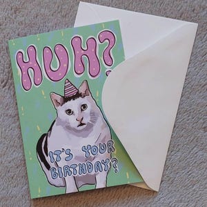 Op de afbeelding: Een groene verjaardagskaart met een witte kat die een roze feestmuts draagt. De kaart zegt "Huh? Is het je verjaardag?" in roze en blauwe letters.