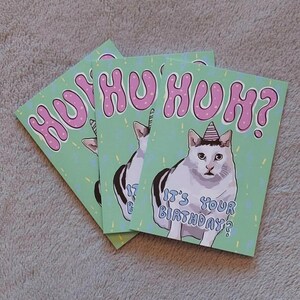 Op de afbeelding: Een groene verjaardagskaart met een witte kat die een roze feestmuts draagt. De kaart zegt "HUHU HUH? Is het je verjaardag?" in roze letters.