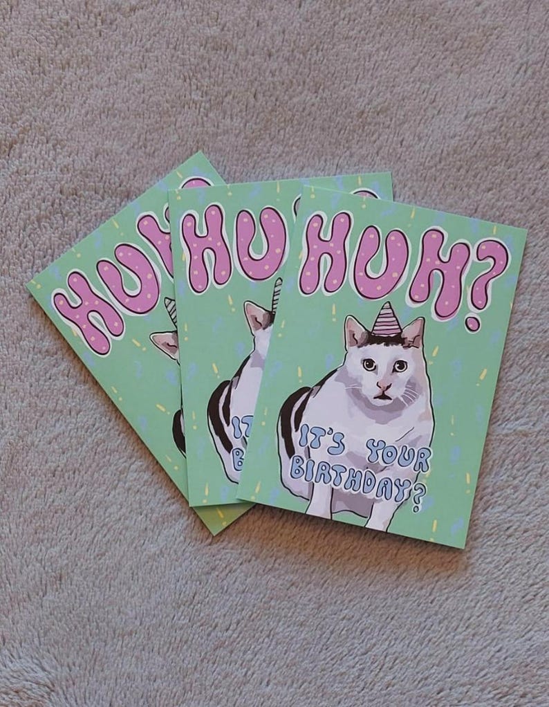 Op de afbeelding: Een groene verjaardagskaart met een witte kat die een roze feestmuts draagt. De kaart zegt "HUHU HUH? Is het je verjaardag?" in roze letters.