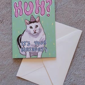 Op de afbeelding: Een groene verjaardagskaart met een witte kat die een roze feestmuts draagt. De kaart zegt "Huh? Is het je verjaardag?" in een kleurrijk lettertype.